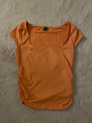 Orange topp med rynkade sidor från Shein - Orange topp från Shein med fyrkantig ringning och korta ärmar. Rynkade sidor och mjukt stretchigt material som sitter skönt. Jätte fin på sommaren, när man är brun. Använd cirka två gånger. Passformen syns på första bilden, med samma tröja fast annan färg.