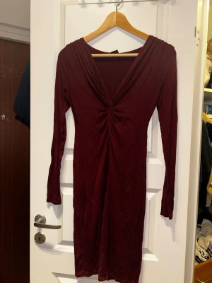 Vinröd långärmad klänning XS - Säljer en vinröd långärmad klänning i storlek XS. Klänningen har en snygg v-ringning med twistad detalj framtill och är figurnära i modellen. Tyget är mjukt och stretchigt, perfekt för dig som gillar en stilren look.