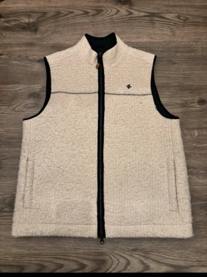 Beige bit  fleeceväst från Morris - Säljer en beige fleeceväst från Morris med svart dragkedja och diskret logga på bröstet. Västen har hög krage, två fickor och svarta detaljer längs kanterna. Perfekt för lager-på-lager och riktigt skön att bära under kyliga dagar.