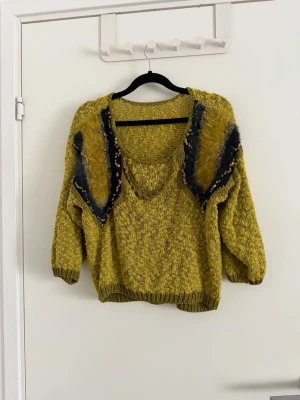 Grön stickad med fluffiga detaljer - Unik grön stickad tröja med coola fluffiga detaljer och pärlor på axlarna. Tröjan har trekvarts ärm och en avslappnad passform. Perfekt för dig som vill sticka ut med en mysig och kreativ look. Aldrig använd. Köpt second hand.