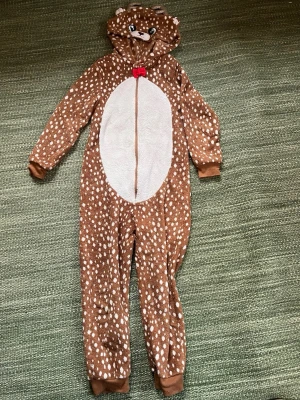 Brun prickig overall från H&M - Supermysig brun Ren-overall från H&M med vita prickar och huva med söta öron och horn. Dragkedja framtill och röd rosett vid halsen. Mjuk fleece på insidan och vita detaljer på magen och svansen. Perfekt för chill dagar eller maskerad.