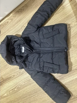Svart pufferjacka barn - Säljer en svart pufferjacka från H&M med huva och dragkedja framtill. Jackan har två stora fickor och är perfekt för kalla dagar. Den har en klassisk, lite boxig passform och är tillverkad i syntetmaterial.