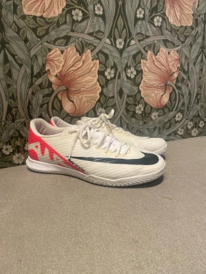 Nike Air Zoom - Säljer ett par Nike Air Zoom fotbollsskor i vitt med svarta detaljer och coola rosa inslag på hälen. Skorna har snörning och en låg profil med platt sula, perfekta för inomhusspel. Tillverkade i syntetmaterial för lätt känsla och bra grepp. Hör gärna av dig vid frågor!
