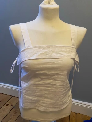 Vit linnetopp med knytband - Säljer en enkel och stilren vit linnetopp med breda axelband och knytband i sidorna. Toppen har rak passform och är tillverkad i ett lätt bomullstyg, perfekt för varma dagar. Passar till jeans, kjol eller shorts för en clean look.. Priset kan diskuteras. Skriv vid frågor😊