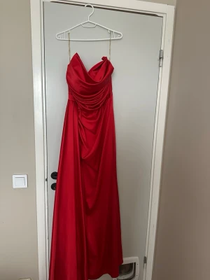 Röd långklänning med drapering - Snygg röd långklänning med tunna axelband och vacker drapering över bysten och magen. Klänningen har en elegant, svepande siluett och är perfekt för speciella tillfällen. Materialet har en lätt glansig finish som ger en lyxig känsla.