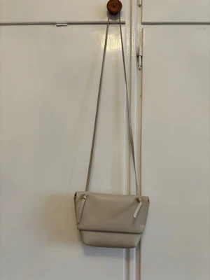Beige axelväska från Acne Studios - Säljer en stilren beige axelväska från Acne Studios i slätt skinn. Väskan har minimalistisk design med vikbar front och knutdetaljer på axelremmen. Perfekt för dig som gillar enkel och modern stil.