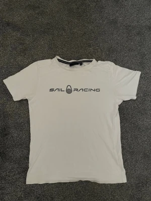 Vit t-shirt från Sail Racing - Vit t-shirt från Sail Racing med svart logga och tryck på bröstet samt en mindre logga i nacken. Klassisk rund halsringning och korta ärmar. Tillverkad i mjuk bomull som känns skön mot huden.