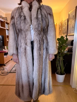 Lång pälsjacka av blåräv från Söderlunds - Exklusiv hellång pälsjacka i grå och beige nyanser, designad av skräddare på Päls Söderlunds. Tillverkad av helryckt blåräv med klassisk krage och raka ärmar. Jackan har en lyxig och tidlös look, perfekt för dig som vill sticka ut med något unikt. Passar personer över 170