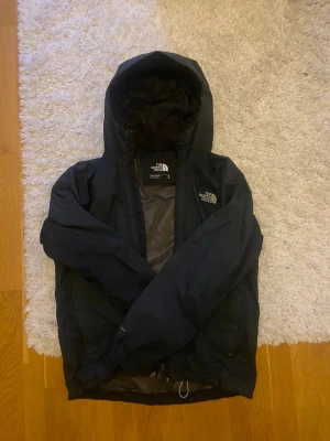 Svart vindjacka från The North Face - Säljer en svart vindjacka från The North Face i storlek M. Jackan har huva, meshfoder och dragkedja framtill. Perfekt för blåsiga dagar och enkel att slänga på sig. Klassisk logga på bröstet och ryggen. Snygg och funktionell design.