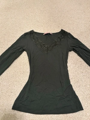 Mörkgrön långärmad topp med spets - Säljer en mörkgrön långärmad topp från Hunkemöller med v-ringning och snygga spetsdetaljer runt halsen. Modellen är figurnära och passar perfekt till jeans eller kjol. Materialet är mjukt och väldigt stretchigt.