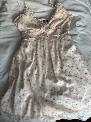 Blommig volangklänning från Vero Moda - Kort klänning från Vero Moda i ljus cremefärg med små rosa blommor. Klänningen har tunna justerbara axelband, volangdetaljer och ett luftigt fall. Storlek (S), skriv via dm vid funderingar 💬🌸