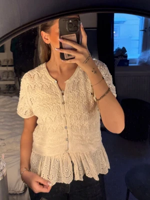 Vit blus från Zara med volang - Supersöt vit blus från Zara med korta ärmar, knappar framtill och vacker spetsstickning. Nederdelen har broderad volangkant som ger en romantisk vibe. Perfekt att styla med jeans eller kjol för en trendig look.