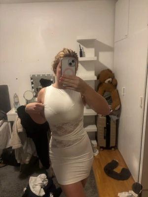 Vit kort klänning med spetsdetaljer - Säljer en vit, tight och ärmlös kortklänning med snygga spetsdetaljer i midjan. Klänningen har en slimmad passform och är tillverkad i ett mjukt syntetmaterial som sitter skönt mot kroppen. Perfekt för dig som gillar stilrena och trendiga plagg med lite extra detaljer.