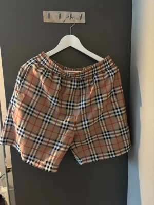  Burberry shorts - Säljer ett par klassiska Burberry shorts i beige med ikoniskt rutigt mönster i svart, vitt och rött. Shortsen har elastisk midja och meshfoder på insidan. Perfekta för sommaren och riktigt snygga till streetwear-looken.