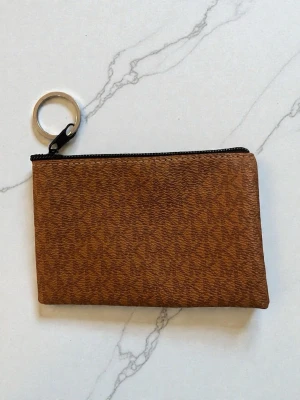 Brun plånbok med nyckelring - Säljer en brun plånbok med diskret mönster och svart dragkedja. Plånboken har en praktisk metallnyckelring i hörnet så du kan fästa den i väskan eller nyckelknippan. Perfekt storlek för kort och småsaker.