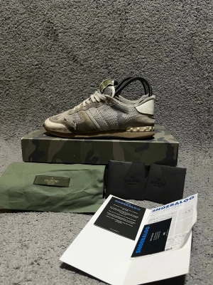Valentino Rockrunner - Säljer ett par Valentino Garavani sneakers i grön/grå camouflage-inspirerad färg. Skorna är 100% äkta och kvitto medföljer 🧾✅ (se bilder). De är slitna och välanvända med synligt slitage på bland annat mesh, mocka och sula, så titta noga på bilderna för att bedöma skicket. Säljs i befintligt skick. Originalkartong, dustbag och Valentino-kort medföljer 📦🔥 Perfekt för dig som vill ha ett par äkta Valentino till ett bättre pris 💸😊