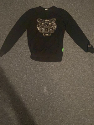 Svart Kenzo sweatshirt med tigertryck - Säljer en svart sweatshirt från Kenzo med ikoniskt tigertryck och Kenzo Paris-brodyr på ärmen. Tröjan har rund hals, långa ärmar och en grön Kenzo-tag i sidan. Perfekt för dig som vill ha en statement piece i garderoben.