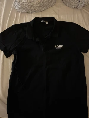 Svart pikétröja från Borg Sportswear - Klassisk svart pikétröja från Borg Sportswear med vit logga på bröstet. Tröjan har krage, korta ärmar och knäppning framtill. Tillverkad i bomull för en skön och avslappnad känsla.