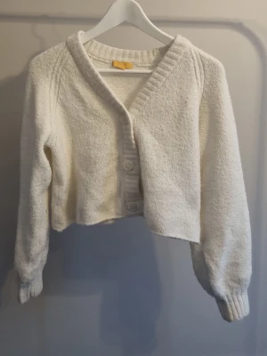 Vit croppad kofta från Gina Tricot - Mysig vit kofta från Gina Tricot i croppad modell. Tröjan har långa ärmar, ribbade muddar och knäppning framtill med tre stora knappar. Perfekt för lager på lager och chill höst- och vinterstil. Perfekt till våren.