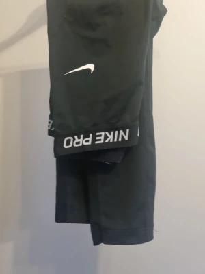 Svarta Nike Pro träningstights - Svarta Nike Pro tights med vit Nike-logga på låret och bred resår i midjan med texten 'NIKE PRO'. Tillverkade i stretchigt syntetmaterial som passar perfekt för träning och sportiga aktiviteter.