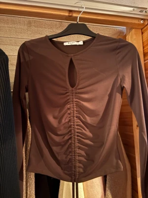 Brun långärmad topp från NA-KD - Säljer en brun långärmad topp från NA-KD med snygg rynkning längs magen och en liten droppformad öppning vid halsen. Toppen har en tight passform och är gjord i ett stretchigt, mjukt material som känns skönt mot huden.