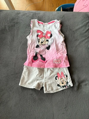Minnie Mouse  pyjamas från H&M - Söt pyjamas från H&M med Minnie Mouse-tryck framtill. Pyjamas är vit och rosa med ärmlös design och har ett stort motiv av Minnie Mouse i rosa klänning och rosett. Tillverkad i mjuk bomull, perfekt för en lekfull och avslappnad stil.