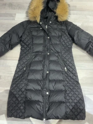 Svart lång dunjacka Rockandblue - Säljer en svart lång dunjacka från Rockandblue med snygg fuskpäls på huvan och quiltade partier på sidorna. Jackan har dragkedja, knappar och två fickor med dragkedja framtill. Perfekt för kalla vinterdagar.