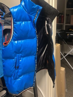 Blå dunväst från Replay - Säljer en riktigt snygg blå dunväst från Replay med hög  och tjock krage som värmer ordentligt. Västen har dragkedja framtill, två fickor med knapp och Replay-logga längs dragkedjan. Detaljer i vitt, rött och svart vid ärmöppning och nederkant. Perfekt för lager-på-lager under höst och vinter.