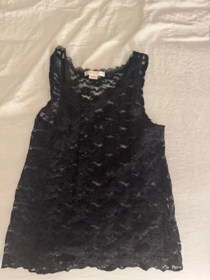 Svart spetslinne från Gina Tricot - Säljer ett svart linne från Gina Tricot i genombruten spets med fina spetskanter vid halsringningen. Linnet är ärmlöst och har en feminin, lätt transparent look som passar perfekt till lager på lager eller som statement-plagg.