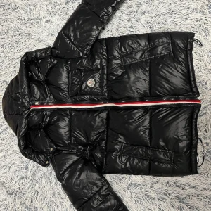 Svart Moncler dunjacka med huva - Svart dunjacka från Moncler med glansig finish, huva och coola fickor. Jackan har en rödvit randig dragkedja och Moncler-logga på bröstet. Insidan har ett serietidningsmönster och flera innerfickor. Perfekt för dig som vill ha en snygg och varm jacka.Montbeliard korta dunjacka för män är en klassisk Moncler-modell och tillverkad av glansigt nylonlack.