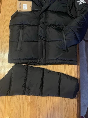 Burbbury jacka *limited stock rabbat* - Säljer en svart pufferjacka från Burberry med avtagbara ärmar och rutigt innerfoder. Jackan har dragkedjor på fickorna, tryckknappar framtill och klassisk Burberry-logga på bröstet. Perfekt för dig som vill ha en varm och stilren jacka med premiumkänsla.