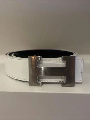 Vit skärp med metallspänne - Snyggt vitt skärp med stort, blankt metallspänne i form av ett H. Skärpet har stilrena sömmar längs kanterna och är gjort i ett slätt material som ger en clean look. Perfekt accessoar för att lyfta vilken outfit som helst.
