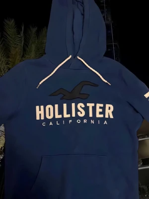Blå hoodie från Hollister med tryck - Blå hoodie från Hollister med stor logga och fågel på bröstet samt text på ena ärmen. Tröjan har huva med vita snören och en stor magficka. Mjuk insida och klassisk passform, perfekt för chill dagar. Litet hål på sidan av armen, påverkar inte användningen och märks knappt av! 