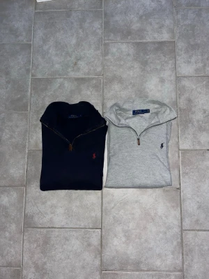 Half zip tröjor från Polo Ralph Lauren - Två snygga half zip tröjor från Polo Ralph Lauren. En marinblå med röd logga och en ljusgrå med marinblå logga. Båda har dragkedja vid halsen och klassisk broderad logga på bröstet. Perfekta för lager på lager under höst och vinter.