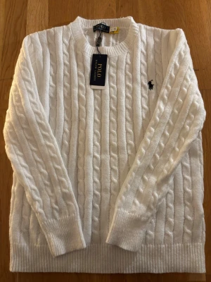 Vit kabelstickad tröja Polo Ralph Lauren - Vit kabelstickad tröja från Polo Ralph Lauren med klassisk rund hals och svart broderad logga på bröstet. Tröjan har ribbade muddar vid ärmslut och nederkant, och är långärmad. Perfekt för dig som gillar stilren och tidlös design.