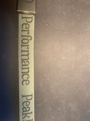 Grönt Peak Performance bälte - Snyggt militärgrönt bälte från Peak Performance med svart metallspänne och tydlig logga längs bandet. Bältet är gjort i slitstark polyester och har en clean, sportig look som passar till jeans eller cargobyxor.