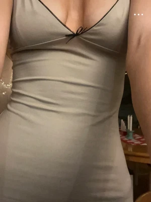 Beige slipklänning från Bershka - Säljer en beige lång klänning från Bershka med tunna axelband och v-ringning. Klänningen har en liten svart rosett framtill och svarta kanter vid bysten. Materialet är glansigt och känns som satin, vilket ger en lyxig känsla.