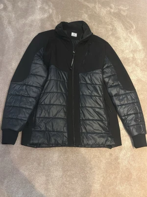 Svart pufferjacka från C.P. Company - Säljer en svart pufferjacka från C.P. Company med quiltad design och dragkedja framtill. Jackan har hög krage, ribbade muddar och flera praktiska fickor. Materialet är polyester och syntet, vilket gör den lätt och bekväm. Perfekt för dig som vill ha en stilren och funktionell jacka.