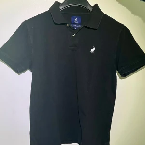 Svart pikétröja från Polo Ralph Lauren - Säljer en klassisk svart pikétröja från Polo Ralph Lauren. Den har en broderad vit logga på bröstet och är i storlek M. Perfekt för en stilren och avslappnad look. Tröjan är i bra skick och redo för nya äventyr!