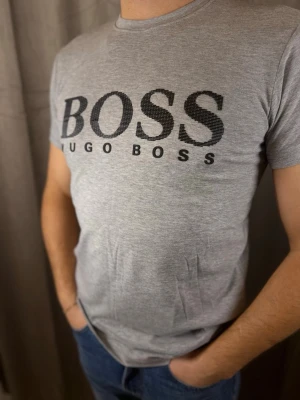 Grå t-shirt från Hugo Boss - Snygg grå t-shirt från Hugo Boss med stor svart logga på bröstet. Klassisk rund halsringning och korta ärmar. Tillverkad i mjuk bomull som känns skön mot huden. Perfekt för dig som gillar stilrena och trendiga plagg med tydlig branding.