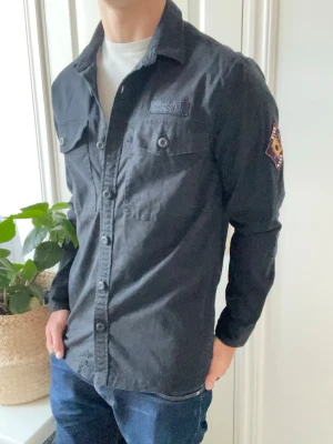 Svart overshirt från Superdry L - Superdry overshirt med loggor på bröstet och axeln. Väldigt snygg och väldigt najs passform! Inga defekter och sittter lite stort på modellen som är 180 cm 75 kg och bär storlek L! Den är igentligen hel svart 