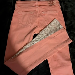 True Religion rosa jeans med spetsdetalj - Säljer ett par ljusrosa jeans från True Religion med snygg spetsdetalj i vitt på insidan av bensluten. Klassisk femficksmodell, silverfärgade knappar och logopatch bak. Även sydda med spets till bootcut. Storlek 27, passar mig som bär ungefär s