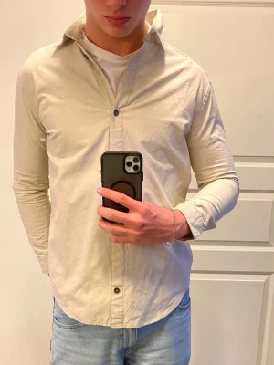Beige skjorta från Timberland - Säljer en stilren beige skjorta från Timberland i relaxed fit. Skjortan är tillverkad i 100% bomull, har klassisk krage, långa ärmar och knappar framtill. Diskret Timberland-logga broderad på bröstet. Perfekt till jeans eller chinos.