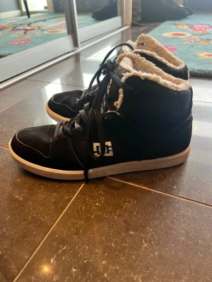Svarta DC high top sneakers med pälsfoder - Säljer ett par svarta DC high top sneakers med vit sula och snörning. Skorna har mjukt fuskpälsfoder på insidan för extra värme och komfort. Ovandelen är i skinn och syntet, perfekt för kalla dagar. Klassisk DC-logga på plösen och yttersulan.