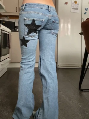 Ljusblå jeans med svarta stjärnor - Unika ljusblå jeans med bootcut passform och två stora svarta stjärnor på bakfickan och benet. Från mitt uf: truestarjeans, Jeansen har tyvärr råa, slitna bensluten pga att de är inköpta begagnat. Storlek 36, måtten: ytterben:104cm innerben:87 lårlängd:17