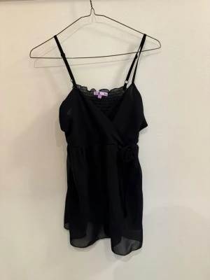 Svart omlott linnetopp från Zoul - Snygg svart omlott linnetopp från Zoul med smala justerbara axelband och dekorativ rosett i midjan. Toppen har transparenta meshdetaljer upptill och en luftig känsla, perfekt för att skapa en chic look.