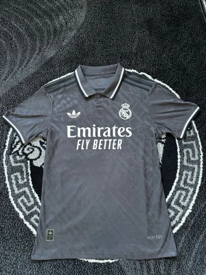 Grå Real Madrid Mbappé tröja Adidas - Säljer en grå Real Madrid fotbollströja från Adidas med Mbappé och nummer 9 på ryggen. Tröjan har broderade detaljer, klassisk krage och vita ränder på axlarna. Materialet är polyester och har ett subtilt mönster i tyget. Perfekt för fotbollsfans!