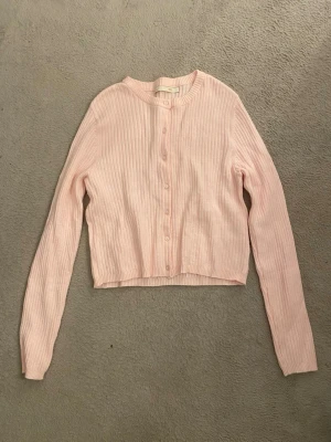 Ljusrosa ribbad kofta - Supersöt ljusrosa kofta med ribbad struktur och knappar framtill. Croppad modell med lång ärm och rund halsringning.