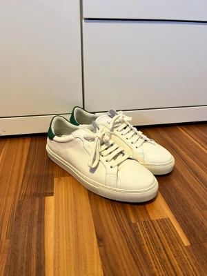 Vita Axel Arigato sneakers med gröna detaljer - Säljer ett par stilrena vita sneakers från Axel Arigato med gröna detaljer på hälen. Skorna har klassisk snörning, rund tå och är tillverkade i slätt skinn. Perfekta för dig som gillar minimalistisk och clean design.