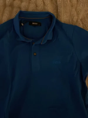 Blå pikétröja från BOSS - Säljer en blå pikétröja från BOSS med klassisk krage och två knappar framtill. Tröjan har diskret BOSS-logga broderad på bröstet och kontrastsöm i kragen. Tillverkad i 100% bomull för en skön känsla.